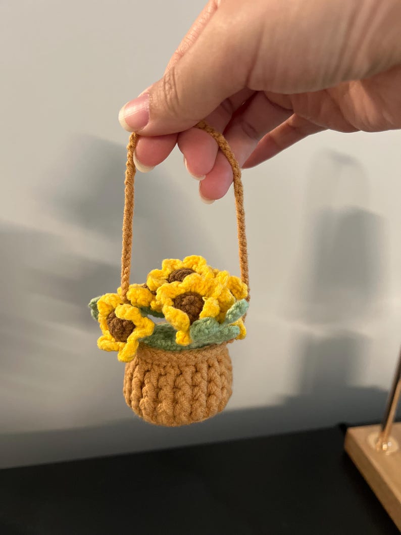 May include: A small, handmade crochet basket featuring a tan base and a matching handle. The basket is filled with three bright yellow sunflowers with brown centers and light green leaves, all crafted from yarn in a detailed, textured stitch.