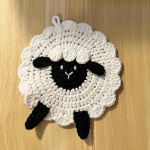 Op de afbeelding: Een handgemaakte gehaakte pannenlap in de vorm van een schaap. De pannenlap is wit met een zwart gezicht, oren en poten. Het heeft een geschulpte rand en een lus om op te hangen. Het schaapontwerp is gecentreerd op een lichtbruin oppervlak.