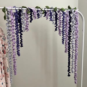 Peut inclure: Une guirlande de glycine au crochet faite &agrave; la main dans les tons de violet, de lavande et de gris, avec des accents de feuilles vertes. La guirlande est suspendue &agrave; une tringle en m&eacute;tal blanc, cr&eacute;ant un affichage floral d&eacute;coratif.