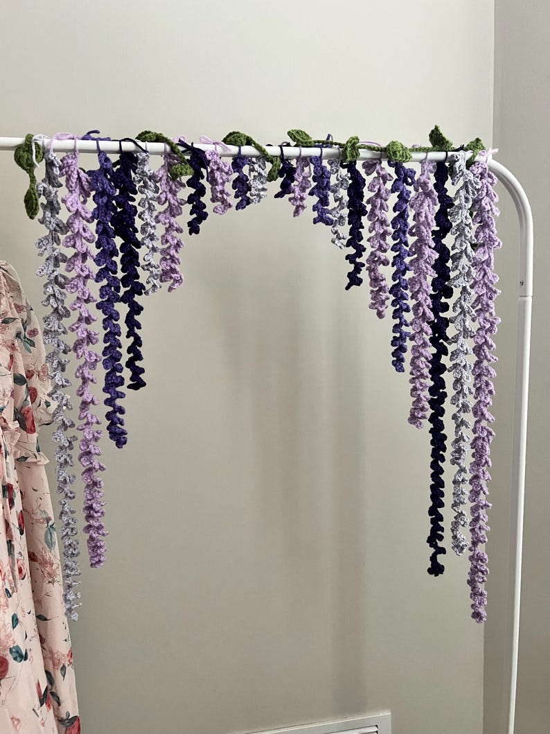 Peut inclure: Une guirlande de glycine au crochet faite &agrave; la main dans les tons de violet, de lavande et de gris, avec des accents de feuilles vertes. La guirlande est suspendue &agrave; une tringle en m&eacute;tal blanc, cr&eacute;ant un affichage floral d&eacute;coratif.