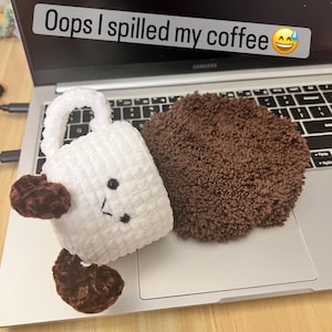 May include: A white crocheted coffee cup with a brown coffee spill, placed on a laptop. The cup has a handle and a smiling face. The text "Oops I spilled my coffee" is above the cup.
