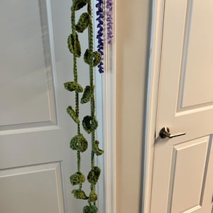 Peut inclure: Une vigne verte crochet&eacute;e &agrave; la main, orn&eacute;e de feuilles et d'accents circulaires, suspendue verticalement. Un brin s&eacute;par&eacute; de fleurs violettes crochet&eacute;es est &eacute;galement visible. La d&eacute;coration est suspendue devant une porte blanche.