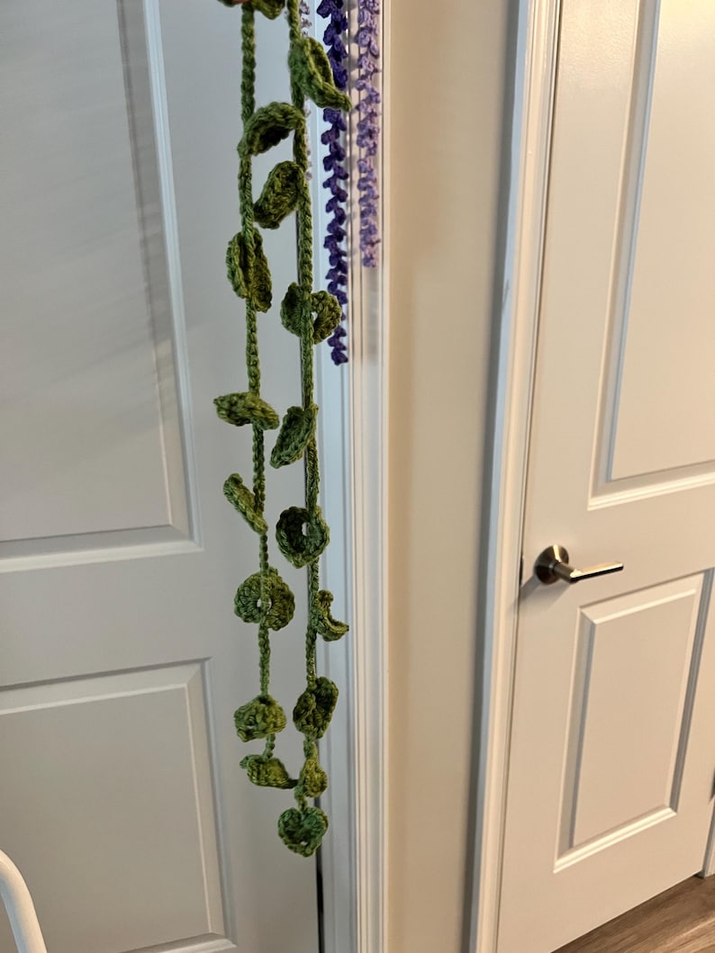 Peut inclure: Une vigne verte crochet&eacute;e &agrave; la main, orn&eacute;e de feuilles et d'accents circulaires, suspendue verticalement. Un brin s&eacute;par&eacute; de fleurs violettes crochet&eacute;es est &eacute;galement visible. La d&eacute;coration est suspendue devant une porte blanche.