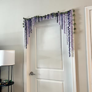 Peut inclure: Une porte blanche est orn&eacute;e d'une guirlande florale. La guirlande pr&eacute;sente des fleurs de type glycine violettes et lavande suspendues avec des feuilles vertes. L'arrangement est mont&eacute; au-dessus du cadre de la porte.