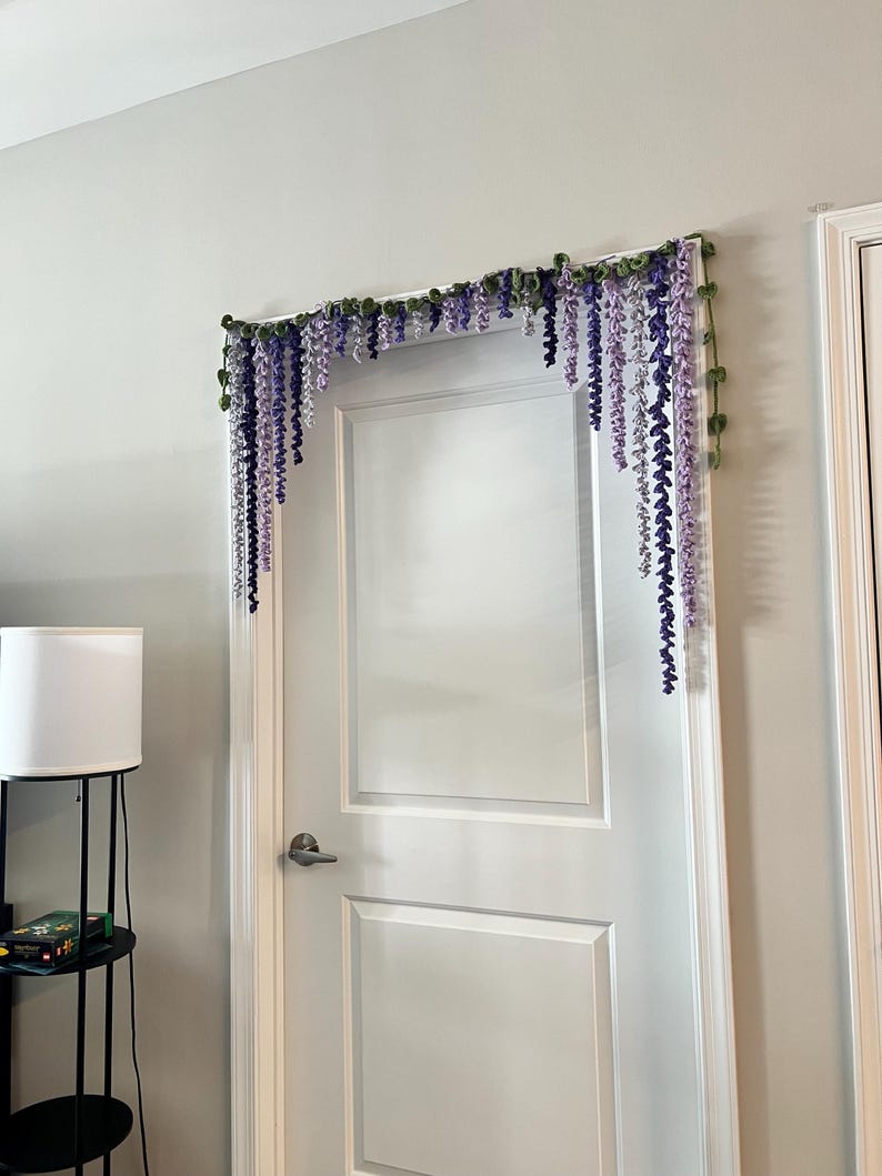 Peut inclure: Une porte blanche est orn&eacute;e d'une guirlande florale. La guirlande pr&eacute;sente des fleurs de type glycine violettes et lavande suspendues avec des feuilles vertes. L'arrangement est mont&eacute; au-dessus du cadre de la porte.