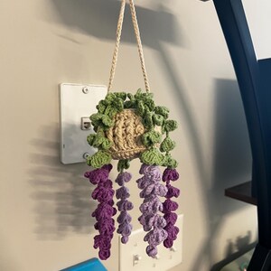 Peut inclure: Un pot de fleurs suspendu au crochet fait &agrave; la main, avec un pot beige, des feuilles vertes et des fleurs de type glycine violettes et lavande. Le pot est suspendu par un cordon beige. Un objet d&eacute;coratif unique pour la d&eacute;coration int&eacute;rieure.