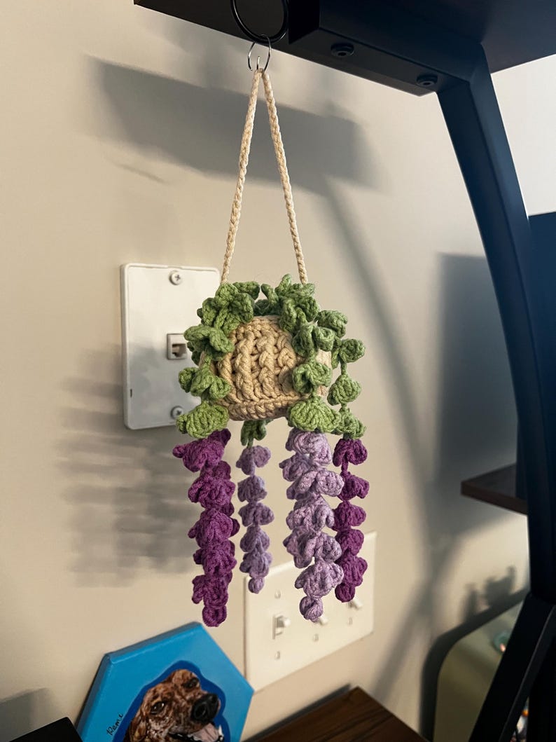 Peut inclure: Un pot de fleurs suspendu au crochet fait &agrave; la main, avec un pot beige, des feuilles vertes et des fleurs de type glycine violettes et lavande. Le pot est suspendu par un cordon beige. Un objet d&eacute;coratif unique pour la d&eacute;coration int&eacute;rieure.