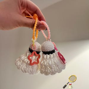 Puede incluir: Dos volantes de bádminton blancos de ganchillo con detalles negros y amuletos de estrella y corazón naranja y rosa. Los volantes están unidos a llaveros naranjas y rosas. Un emoji de raqueta y volante de bádminton está en la esquina inferior derecha.