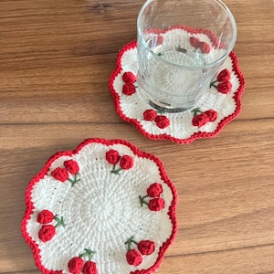 Peut inclure: Deux dessous-de-verre en crochet blanc avec une bordure rouge et des motifs de cerises rouges. Les dessous-de-verre sont sur une surface en bois. Un dessous-de-verre a un verre transparent sur le dessus.