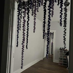 Peut inclure: Une porte est orn&eacute;e de guirlandes de glycine crochet&eacute;es suspendues. Les vignes tombent en d&eacute;grad&eacute;s de violet et de gris, cr&eacute;ant une d&eacute;coration florale fantaisiste. La pi&egrave;ce au-del&agrave; est visible &agrave; travers la porte.