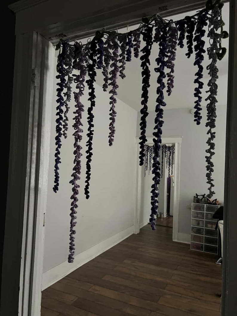 Peut inclure: Une porte est orn&eacute;e de guirlandes de glycine crochet&eacute;es suspendues. Les vignes tombent en d&eacute;grad&eacute;s de violet et de gris, cr&eacute;ant une d&eacute;coration florale fantaisiste. La pi&egrave;ce au-del&agrave; est visible &agrave; travers la porte.