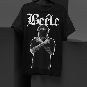 Pode incluir: Camiseta preta com a palavra "Beéle" em uma fonte estilizada. A camiseta apresenta um gráfico em preto e branco de uma pessoa com os braços cruzados, tatuagens e uma cobertura facial. A camiseta é exibida em um cabide preto.