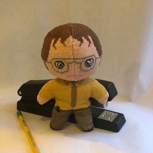 May include: A brown and yellow plush toy of a character wearing glasses. The toy is sitting on a black stapler.