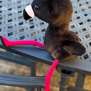 Peut inclure: Un oiseau en peluche avec un corps, une tête et des ailes noirs, un bec blanc et rose, et des pattes et des pieds rose vif. L'animal en peluche est posé sur une surface grise.