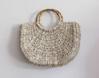 May include: A semi-circular woven straw handbag featuring a light beige natural fiber construction with a tight, vertical weave pattern. The bag includes two rounded, segmented bamboo handles attached to the top edge of the woven body.