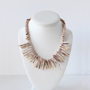 May include: A necklace composed of numerous thin, elongated, natural shell fragments in shades of cream, tan, and dark brown. The shells are arranged in a dense, layered fringe pattern around the neckline, creating a textured, multi-dimensional appearance.