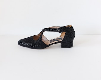 May include: Black, textured, low-heeled shoes with a T-strap design. The shoes have a pointed toe and a small buckle closure. The heels are approximately 2 inches high. The shoes are likely vintage or retro style.