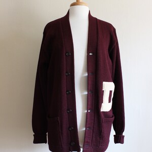 May include: A maroon cardigan sweater with a white letter "D" patch on the left side. The sweater has a button-up front, two front pockets, and ribbed cuffs. The sweater is displayed on a mannequin.