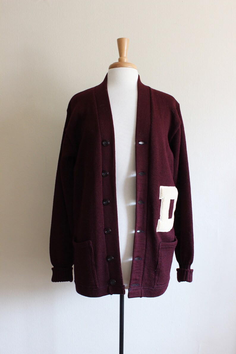 May include: A maroon cardigan sweater with a white letter "D" patch on the left side. The sweater has a button-up front, two front pockets, and ribbed cuffs. The sweater is displayed on a mannequin.