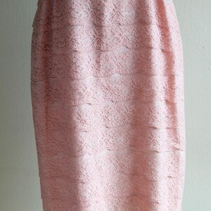 May include: A knee-length dress in a soft pastel pink hue, featuring a layered lace overlay with a floral pattern. The garment is accented at the waist with a smooth, satin-finish bow in a matching pink tone, creating a textured appearance.