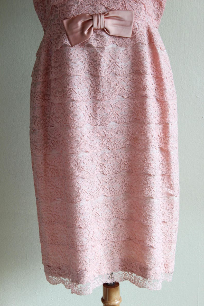 May include: A knee-length dress in a soft pastel pink hue, featuring a layered lace overlay with a floral pattern. The garment is accented at the waist with a smooth, satin-finish bow in a matching pink tone, creating a textured appearance.