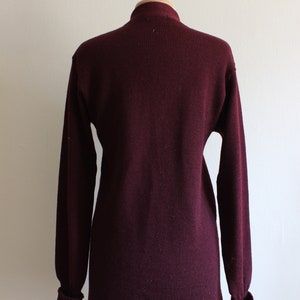 May include: A long-sleeved, burgundy sweater with a high neckline, displayed on a mannequin. The sweater has a ribbed texture and fitted cuffs. The garment is a solid color and appears to be made of a knit material.