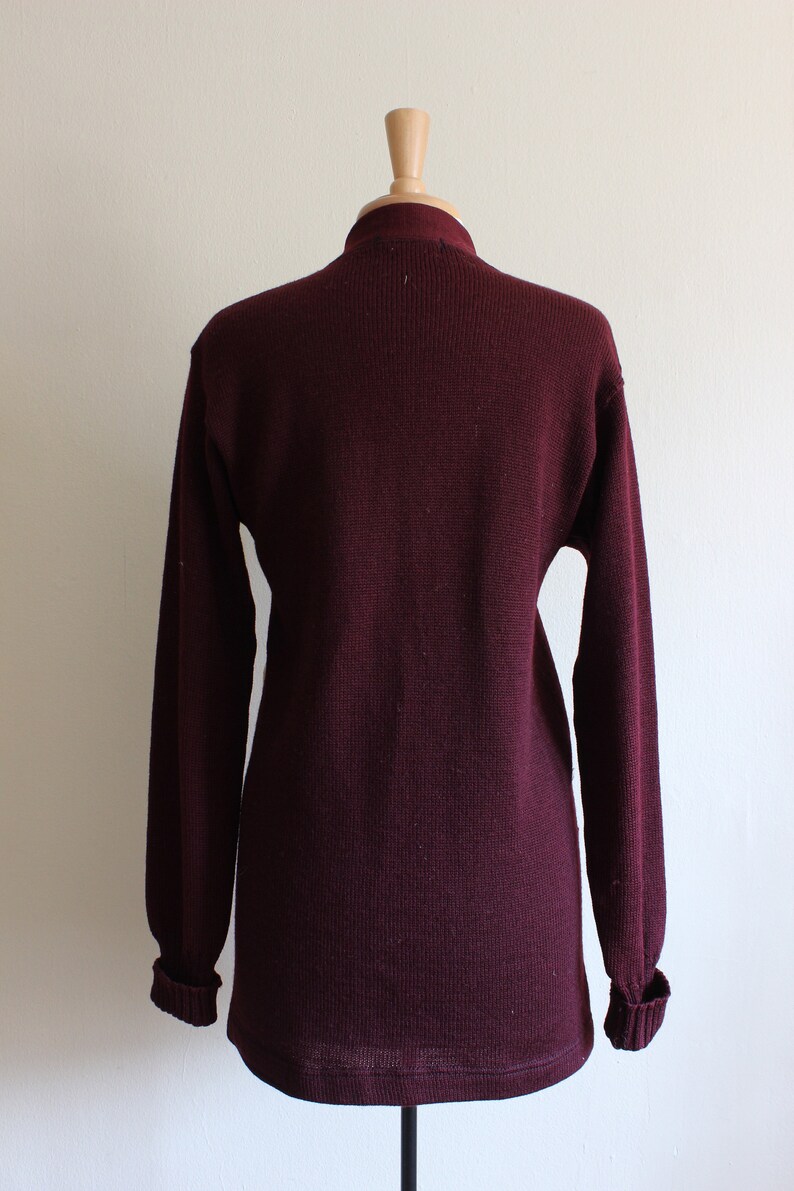 May include: A long-sleeved, burgundy sweater with a high neckline, displayed on a mannequin. The sweater has a ribbed texture and fitted cuffs. The garment is a solid color and appears to be made of a knit material.
