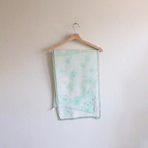May include: A light green silk scarf with a floral pattern. The scarf is hanging on a wooden hanger.