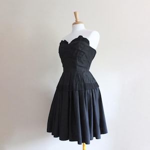 May include: A strapless black dress displayed on a mannequin. The dress features a fitted bodice with a sweetheart neckline and a pleated skirt that flares out. The dress is made of a shiny fabric.