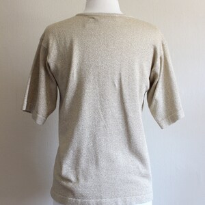 May include: A gold-toned, short-sleeved knit top on a mannequin. The top has a round neckline and a subtle shimmer. The fabric appears to be a soft knit material, suitable for casual wear. The mannequin is white.