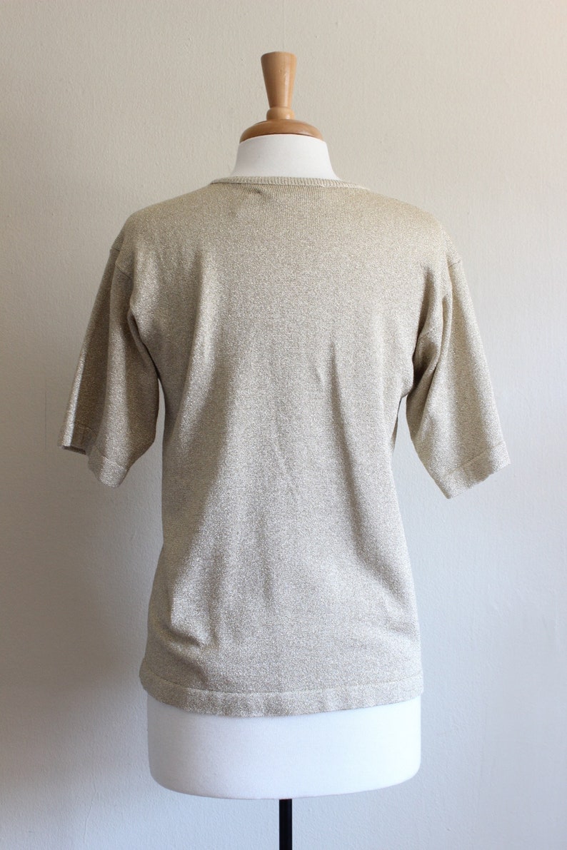 May include: A gold-toned, short-sleeved knit top on a mannequin. The top has a round neckline and a subtle shimmer. The fabric appears to be a soft knit material, suitable for casual wear. The mannequin is white.