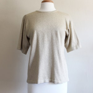 May include: A gold-coloured, short-sleeved knit top on a mannequin. The top features a round neckline and a textured finish. The fabric has a subtle shimmer, giving it a luxurious appearance.