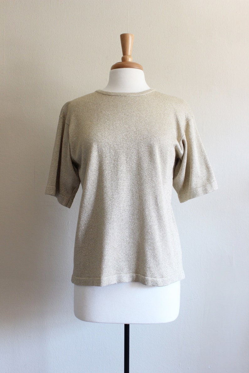 May include: A gold-coloured, short-sleeved knit top on a mannequin. The top features a round neckline and a textured finish. The fabric has a subtle shimmer, giving it a luxurious appearance.