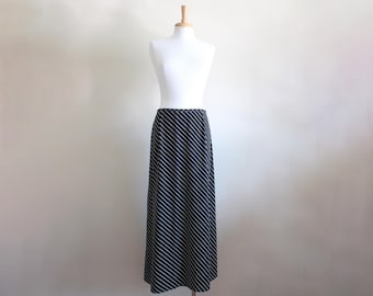 May include: A floor-length black skirt featuring a repeating pattern of thin, diagonal white stripes. The skirt has a straight, streamlined silhouette and is displayed on a white mannequin torso, showcasing the full length of the garment.