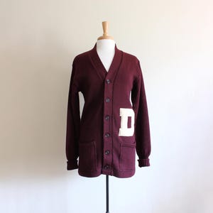 May include: A maroon button-down cardigan sweater with a white letter "D" patch on the front. The sweater has a V-neck, long sleeves, and two front pockets. The sweater is displayed on a mannequin.