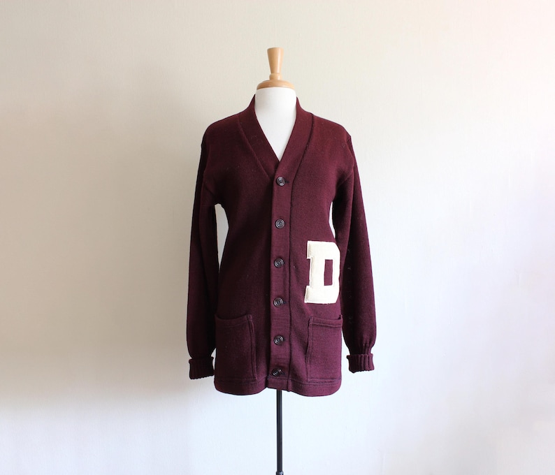 May include: A maroon button-down cardigan sweater with a white letter "D" patch on the front. The sweater has a V-neck, long sleeves, and two front pockets. The sweater is displayed on a mannequin.