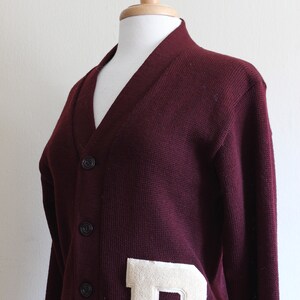 May include: A maroon V-neck cardigan sweater with a large, off-white felt letter "D" patch. The sweater has a button-up front and is displayed on a mannequin. The knitwear is a classic style.