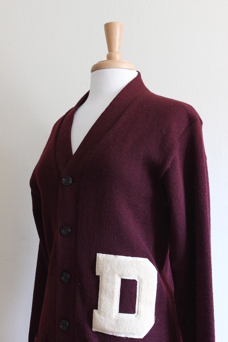 May include: A maroon V-neck cardigan sweater with a large, off-white felt letter "D" patch. The sweater has a button-up front and is displayed on a mannequin. The knitwear is a classic style.