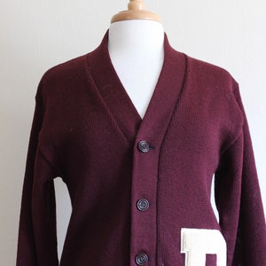 May include: A maroon cardigan sweater with a V-neck and button closure. A large, off-white felt letter "D" is sewn onto the front. The sweater is displayed on a mannequin.