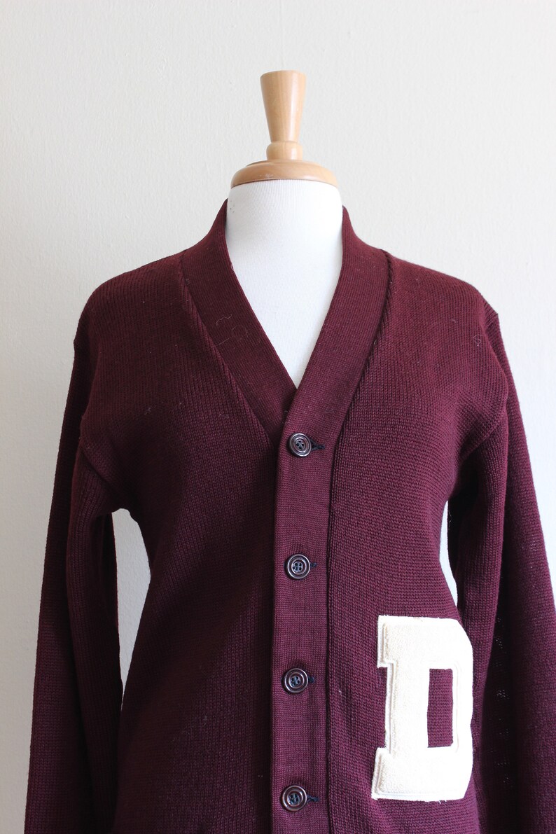 May include: A maroon cardigan sweater with a V-neck and button closure. A large, off-white felt letter "D" is sewn onto the front. The sweater is displayed on a mannequin.