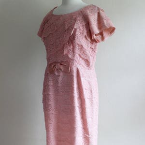 May include: A dusty pink dress featuring an all-over lace overlay with horizontal tiered ruffles throughout the bodice and skirt. The garment has short flutter sleeves, a round neckline, and a small satin bow detail centered at the waistline.