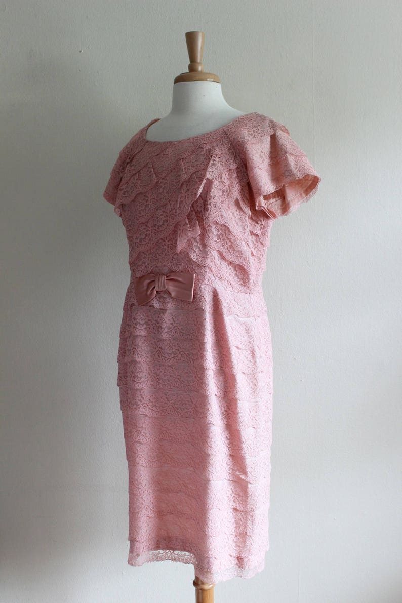 May include: A dusty pink dress featuring an all-over lace overlay with horizontal tiered ruffles throughout the bodice and skirt. The garment has short flutter sleeves, a round neckline, and a small satin bow detail centered at the waistline.