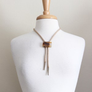May include: A beige bolo tie with a brown and white agate stone pendant. The tie is on a white mannequin.