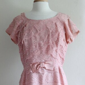 May include: A pale pink dress featuring an all-over floral lace overlay with tiered ruffled sleeves and a matching satin bow detail at the waist. The garment has a rounded neckline and a layered skirt construction, displayed on a mannequin.