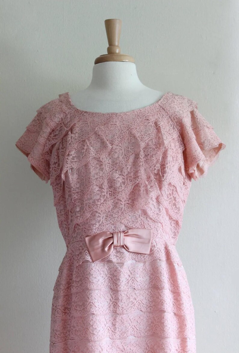 May include: A pale pink dress featuring an all-over floral lace overlay with tiered ruffled sleeves and a matching satin bow detail at the waist. The garment has a rounded neckline and a layered skirt construction, displayed on a mannequin.