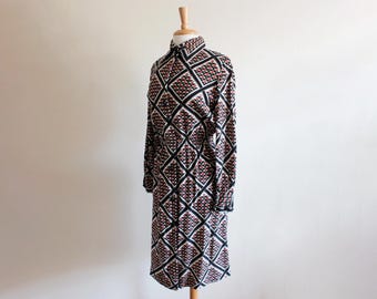 May include: A long-sleeved dress with a geometric pattern in black, white, and red. The dress is displayed on a wooden dress form. The pattern consists of diamond shapes. The dress has a collared neckline and buttoned cuffs.