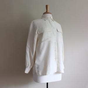 May include: Cream-colored button-down shirt with two chest pockets and long sleeves. The shirt is displayed on a mannequin. The fabric appears to be lightweight and has a subtle sheen. The collar is a classic point style.