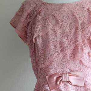 May include: A dusty pink lace dress featuring a round neckline, short layered sleeves, and a horizontal tiered design. The bodice is accented with a smooth, satin-finish bow at the waistline, and the entire garment is crafted from floral lace.