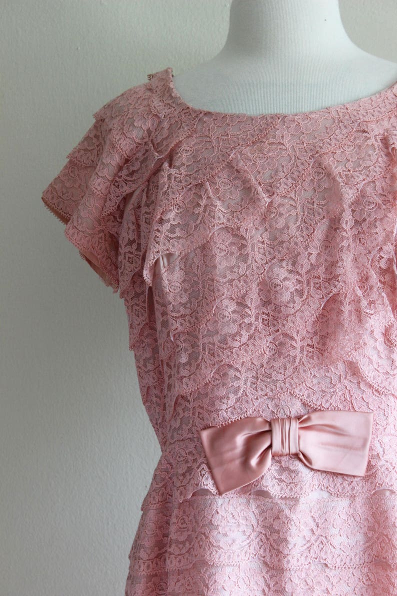 May include: A dusty pink lace dress featuring a round neckline, short layered sleeves, and a horizontal tiered design. The bodice is accented with a smooth, satin-finish bow at the waistline, and the entire garment is crafted from floral lace.