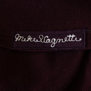 May include: Close-up of a dark burgundy knit fabric with a sewn-on label. The label is a darker shade of burgundy and features the embroidered text "Mike Magnetti" in white thread. The fabric appears to be a sweater or similar garment.